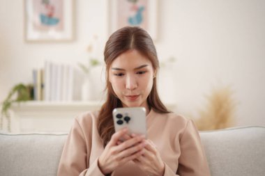 Güzel Asyalı kadın yumuşak bir kanepede rahat bir şekilde otururken akıllı telefonuna bakıyor..