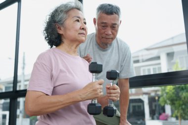 Asyalı son sınıf çifti spor salonunda birlikte çalışıyor, esneme ve güç egzersizlerine odaklanıyor. Yaşlı kadın dambılı tutarken kocası onu cesaretlendiriyor ve sağlıklı bir yaşam tarzı sergiliyor..