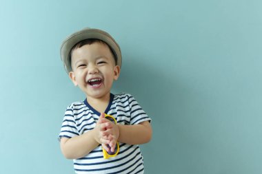 Çizgili tişört ve şık bir şapka giyen neşeli genç Asyalı çocuk mavi izole arka planda ellerini çırparken neşeyle gülüyor, mutluluk ve heyecan saçıyor..