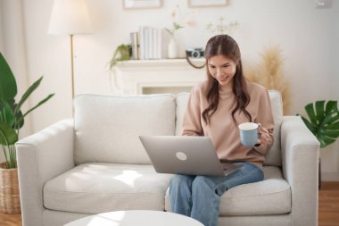 Asyalı genç bir kadın rahat bir oturma odasında kanepede oturmuş elinde bir fincan kahve tutarken dizüstü bilgisayarıyla gülümsüyor..