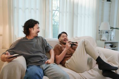 Rahat Asyalı erkekler kanepede birlikte oyun oynuyorlar. İki arkadaş gülümsüyor ve hafta sonu akıllı telefondan arkadaşça bir yarışmanın keyfini çıkarıyorlar..