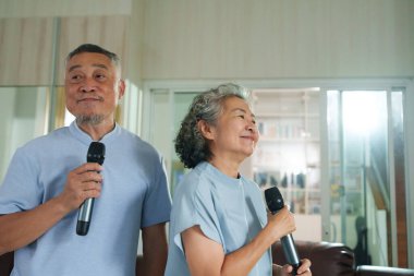 Asyalı yaşlı çift evde neşeyle karaoke yapıyor, mikrofon tutuyor ve gülümsüyor. Parlak ve neşeli bir atmosferi olan rahat bir oturma odasında duruyorlar.