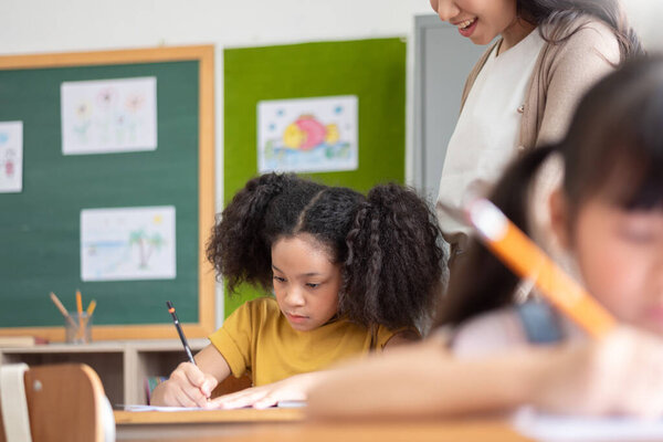 African American girl with curly hair focuses intently on her writing at classroom desk, while teacher offers guidance. classroom and filled with drawings.