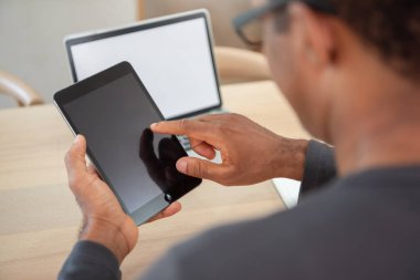 Afrika kökenli Amerikalı bir adam bilgisayarla masada otururken dijital tablet kullanıyor, modern çalışma alanındaki teknoloji ve iletişim üzerine çalışıyor.