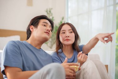 Sıradan mavi tişörtler giyen Asyalı genç çift televizyonda bir şey izliyor. Kadın işaret ediyor ve ikisi de evde meşgul ve rahat..