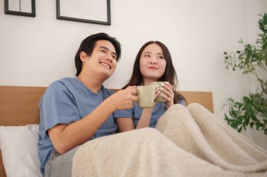 Asyalı genç çift sıcak bir sabah geçirir, sıcak içecekleri yudumlar ve battaniyeye sarınır, sıcak ve rahat bir atmosfer yaratır.