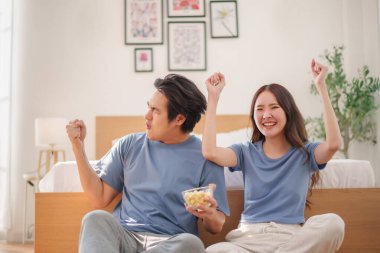 Neşeli Asyalı genç çift gündelik kıyafetleriyle yerde oturup kollarını kaldırarak kutlama yapıyor ve yatak odasında eğleniyorlar.