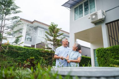 Yeşillik bahçede neşeyle çiçek sulayan Asyalı yaşlı çift modern evin önünde yan yana duruyorlar. Sahne mutluluk ve arkadaşlık anını yakalar.