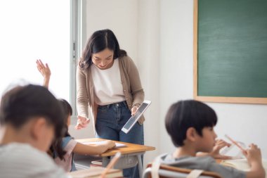 Asyalı Öğretmen dijital tablet kullanan öğrencilerle etkileşime girerken aynı zamanda bir sınıfa katılmayı teşvik eder.