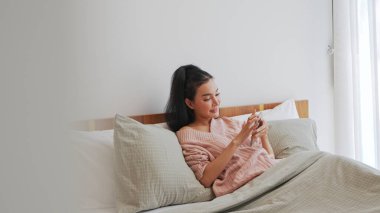 Pembe kazak giyen genç Asyalı kadın yatak odasında yumuşak bir yatak örtüsüyle akıllı telefonunu karıştırmaktan hoşlanıyor..