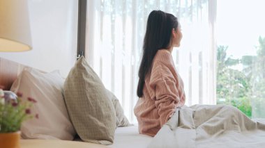 Asyalı bir kadın yatakta oturur, sakin bir açık hava sahnesine pencereden bakar, sabah huzurunun tadını çıkarır..