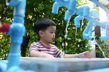 Asyalı çocuk, yeşilliklerle çevrili canlı bir açık hava oyun alanında akan suyla eğlenceli bir keşfe çıkıyor..