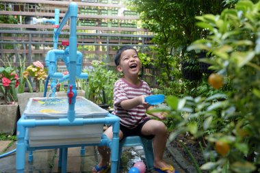 Neşeli Asyalı çocuk bahçede suyla oynuyor, sandalyede oturuyor, dışarıda bir kase tutarken coşkuyla sıçrıyor ve gülüyor..