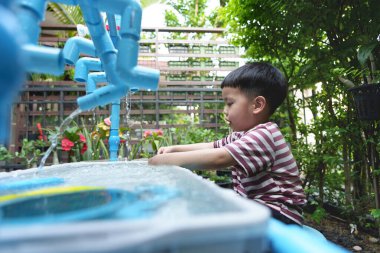 Genç Asyalı çocuk bahçedeki plastik borulardan akan suyla neşeyle oynuyor.