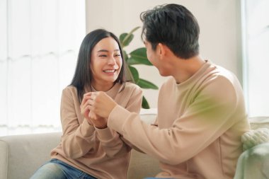 Heyecanlı Asyalı çift birbirlerine gülümsüyor ve keyifli hamilelik testi sonuçlarını oturma odasında kutluyorlar..