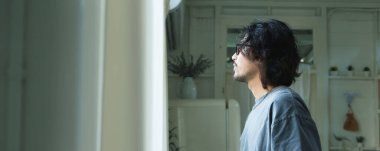 Asyalı bir adam pencerenin yanında durmuş, bir evde derin düşüncelere dalmış bir ifadeyle dışarı bakıyor..