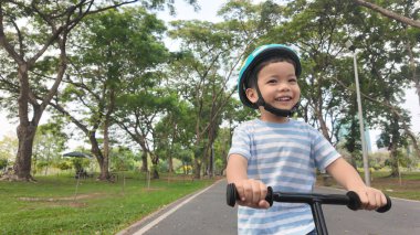 Asyalı çocuk motosiklet sürerken gülümsüyor ya da arka planda ağaçlar olan yeşil bir parkta dengeli bisiklet sürüyor..