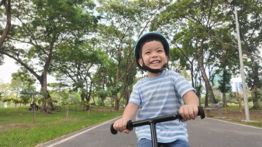 Ağaçlarla ve yeşilliklerle dolu canlı bir parkta dengeli bisiklete binen mutlu Asyalı çocuk açık hava aktivitelerinin neşesini sergiliyor..