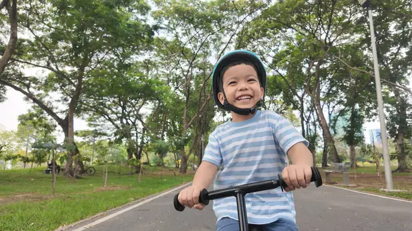 Ağaçlarla ve yeşilliklerle dolu canlı bir parkta dengeli bisiklete binen mutlu Asyalı çocuk açık hava aktivitelerinin neşesini sergiliyor..
