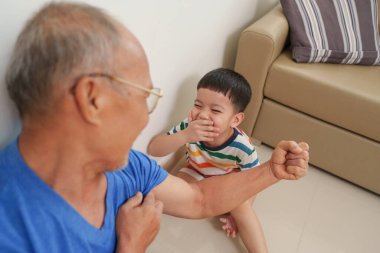 Asyalı çocuk büyükbabasıyla eğlenirken gülüyor, kıdemli adam evde oynaşırken kaslarını gösteriyor..