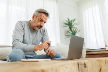 Odaklanmış, gri saçlı, olgun bir adam bir kanepede oturup dizüstü bilgisayarla çalışıyor ve elinde kalemle ekrana bakıyor..