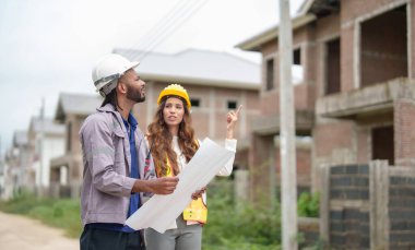 Güvenlik kaskı takan iki inşaat işçisi inşaat projesini konuşurken inşaat alanının planlarını tutuyorlar. Olay yeri incelemeleri takım çalışması ve planlama