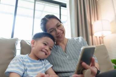 Mutlu Asyalı anne ve küçük oğlu birlikte akıllı telefona bakarken kanepede oturup gülümsüyorlar..