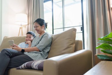 Asyalı anne ve oğlu kanepede rahat bir şekilde otururlar. Oturma odasında birlikte akıllı telefona bakarken gülümserler..