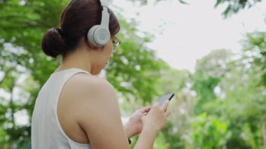 Asian young woman wearing headphones stands in a lush park, immersed in her smartphone. She enjoys a peaceful moment in nature, surrounded by greenery, during a sunny day.