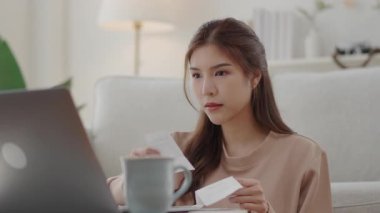 Asyalı kadın oturma odasında, evinde dizüstü bilgisayarla kahve masasında oturmuş finansal belgeleri yoğun bir şekilde analiz ediyor..