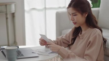 Asyalı bir kadın, oturma odasında dizüstü bilgisayar ve kupayla çevrili bir kahve masasında mali evrakları incelerken stresli hissediyor..