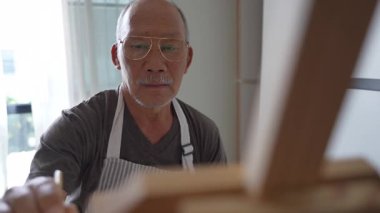 Önlük takan yaşlı bir Asyalı gün içinde evinde tuvale resim çiziyor. Emeklilik sanatsal hobisinin tadını çıkarıyor..