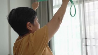 Asyalı üst düzey bir kadın, sakin bir ortamda direnç bantlarıyla gerinmeye ve egzersiz yapmaya odaklanarak evde günlük aktivitelere katılıyor..