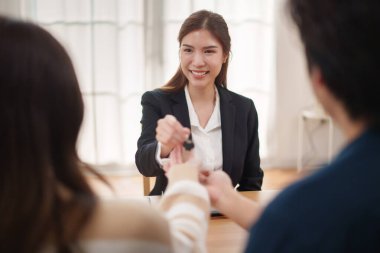 Gülümseyen Asyalı kadın ajan masadaki genç çifte ev anahtarını veriyor, başarılı emlak alışverişi, ev sahipliği ve müşteri memnuniyeti..