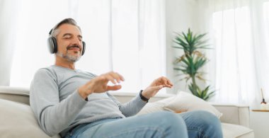 Gülümseyen orta yaşlı kulaklıklı adam evde rahat rahat müzik dinliyor..