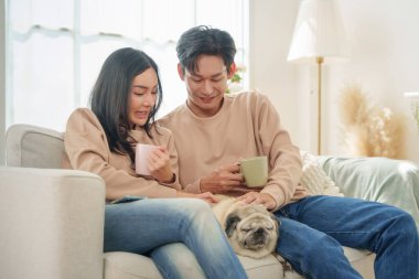 Mutlu Asyalı çift kanepede birlikte kahvenin keyfini çıkarıyorlar. Yanında sevimli köpekleri dinleniyor, gülümsüyor ve içeride dinleniyorlar..