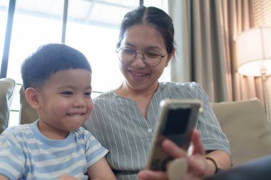 Gözlüklü gülümseyen Asyalı anne ve genç çocuk rahat oturma odasında cep telefonundan eğlenceli bir şey izliyorlar..