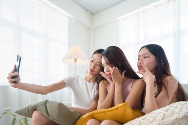 Bir grup Asyalı kız arkadaş kanepede oturup, gülücükler ve eğlenceli pozlarla dolu akıllı telefonlarıyla rahatça selfie çekiyorlar..