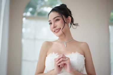 Güzel bir Asyalı gelin içeride şık bir gelinlik içinde poz veriyor. Stüdyoda poz verirken pencereden dışarı bakıp gülümseyen ve mutluluğunu gösteren bir kadın..