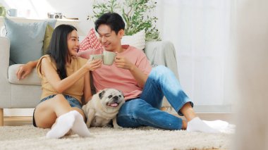 Genç Asyalı çift oturma odasında sevimli pug köpeğiyle oynuyor, birlikte neşeli anları paylaşıyorlar, bağ kurma ve arkadaşlık kavramı.