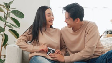 Beyaz koltukta oturan Asyalı çift birbirlerine gülümserken ultrason fotoğrafı çekiyorlar. Hamilelik, neşe ve gelecekteki aile bağları kavramı.