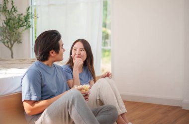 Genç Asyalı erkek ve kadın yerde patlamış mısır paylaşıyor, gülümsüyor ve abur cubur saatinde kaynaşıyorlar.