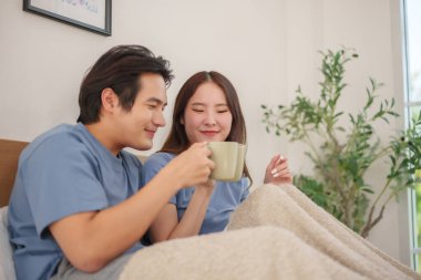Genç Asyalı çift sıcak bir battaniye ve gülümsemeyle yatakta kahvelerini yudumluyor.