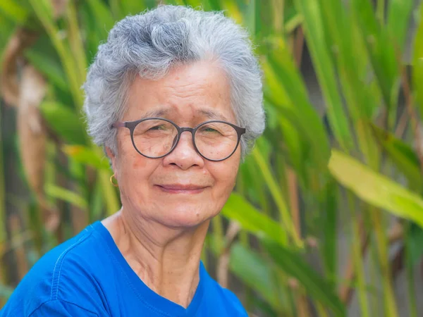 stock image Cheerful senior woman with short gray hair wearing glasses, smiling and looking at the camera while standing in a garden. Space for text. Concept of aged people and healthcare.