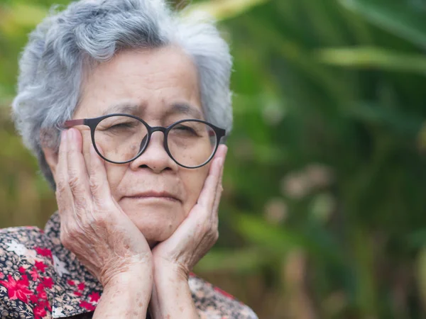 stock image Senior woman with short gray hair, wearing glasses and toothache while standing in a garden. Space for text. Selective focus. Concept of aged people and healthcare.