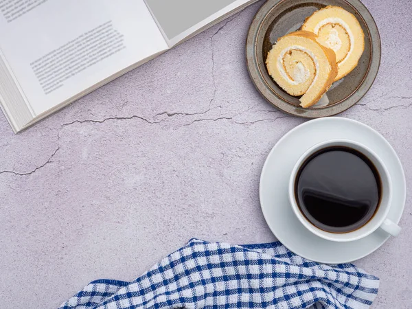 Top view of a book with roll cakes on a plate, a white coffee cup, and a cloth placed on a gray stone background. Space for text. Concept of relaxation
