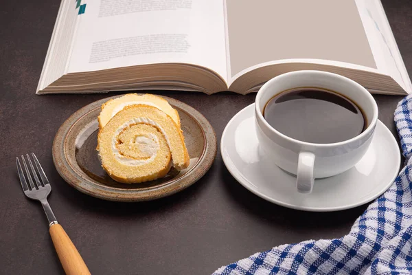 Top view of a book with roll cakes on a plate, a white coffee cup, and a cloth placed on dark gray stone background. Space for text. Concept of relaxation.