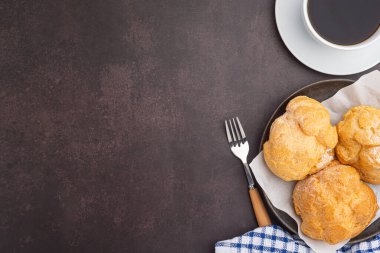Tabaktaki choux kremasının üst görüntüsü ve koyu kahverengi arka planda beyaz bir kahve fincanı. Mesaj için yer var. Rahatlama kavramı.