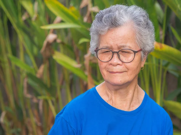 Portrait of a senior woman with short gray hair wearing glasses and looking down while standing in a garden. Space for text. Concept of aged people and healthcare.