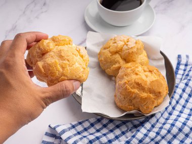 Hand holding a choux cream with choux cream on a plate and a white coffee cup with a cloth placed on a marble table. Space for text. Concept of dessert, beverage and relaxation.